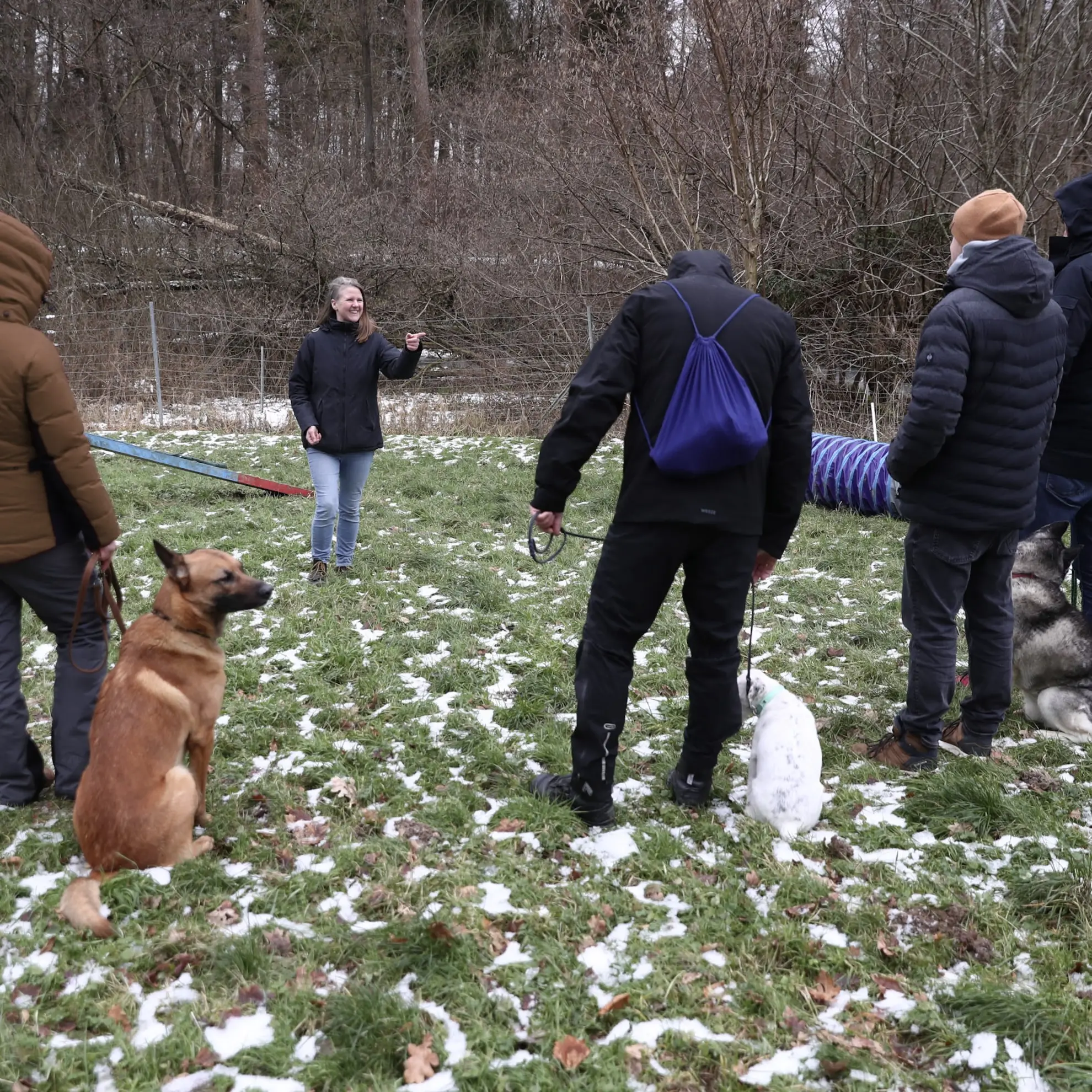 Eine Hundetrainerin steht vor einer Gruppe mit Hunden, zeigt mit dem Finger auf eine Person und lächelt.