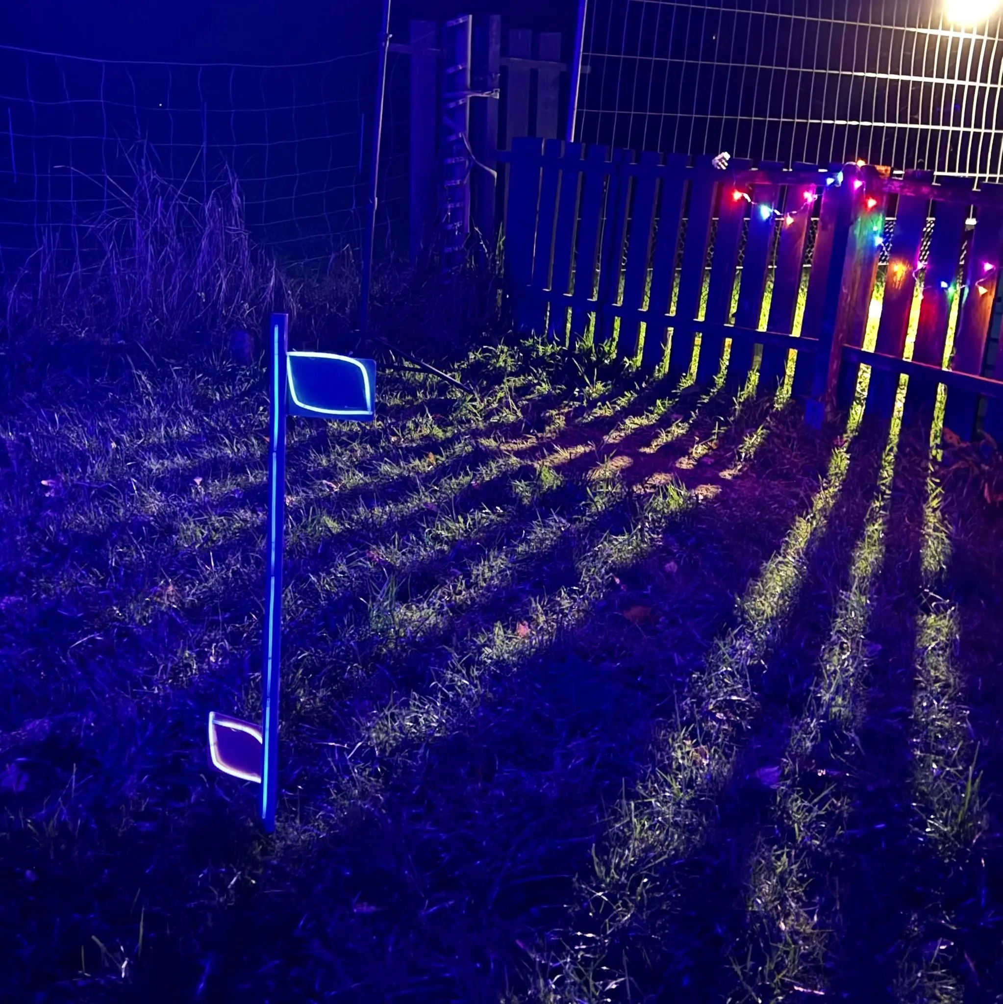 Curvingstange mit Knicklichtern steht vorn einem Zaun mit einer bunten Lichterkette