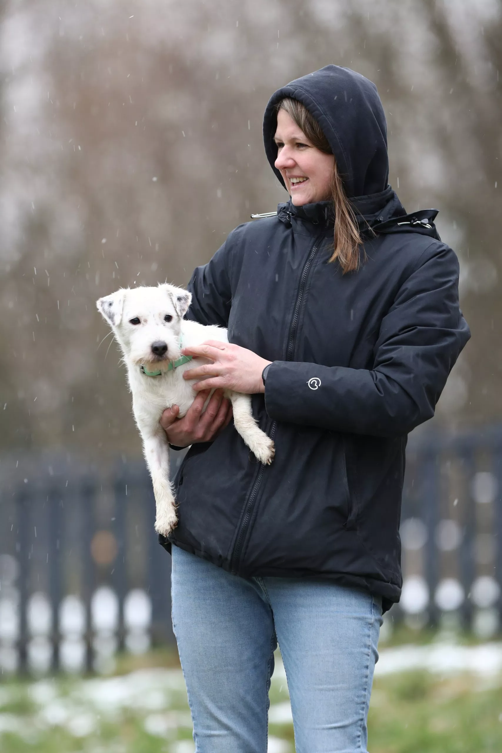 Frau in einer schwarzen Jacke mit Kapuze auf hält einen weißen kleinen Hund auf dem Arm